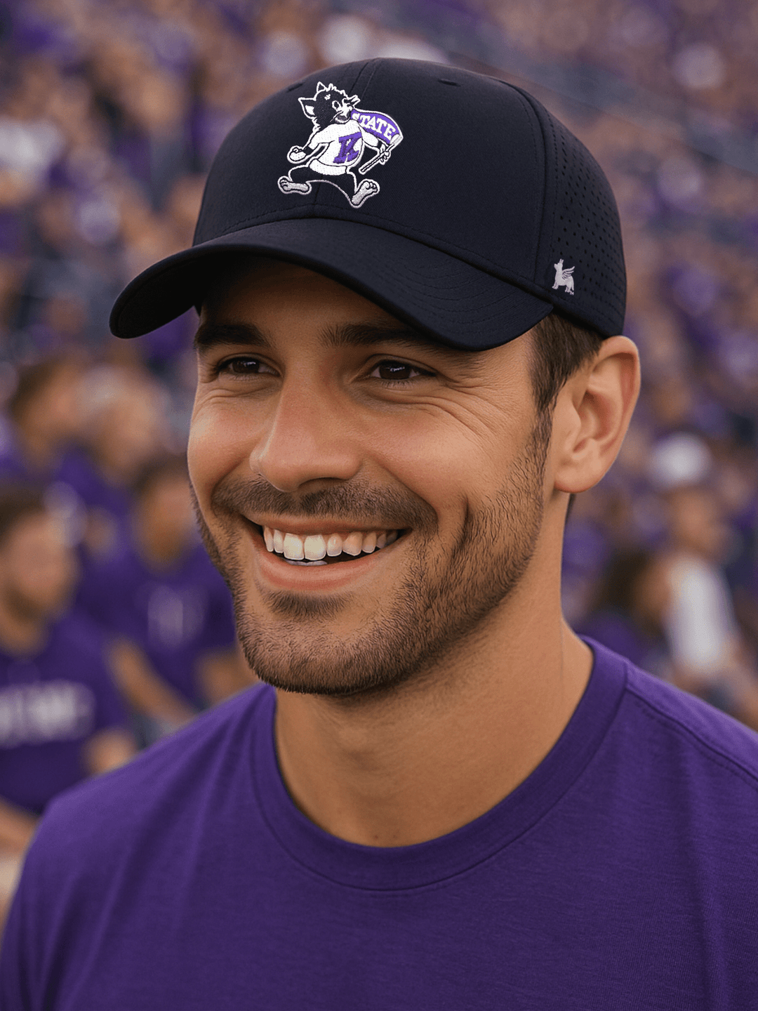 Smiling man in his 30s wearing a black low-profile Kansas State University performance hat with the vintage Willie the Wildcat logo, standing in the crowd at a football game with fans dressed in purple spirit wear