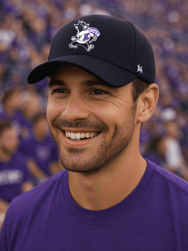 Smiling man in his 30s wearing a black low-profile Kansas State University performance hat with the vintage Willie the Wildcat logo, standing in the crowd at a football game with fans dressed in purple spirit wear