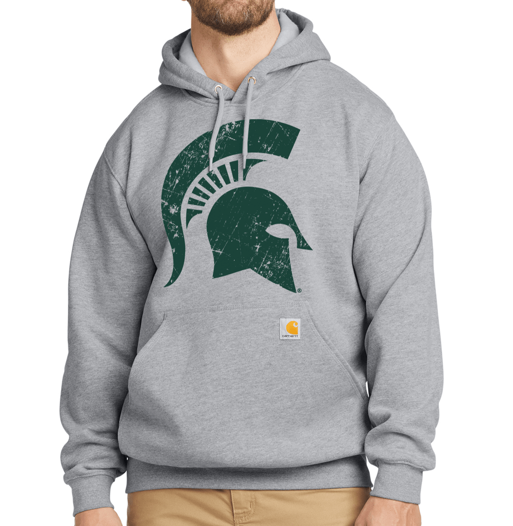 Bearded male model wearing heather grey Carhartt pullover hoodie with large green distressed Spartan helmet print on the chest and Carhartt brand label on the front pocket.