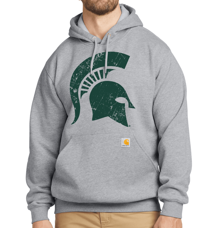 Bearded male model wearing heather grey Carhartt pullover hoodie with large green distressed Spartan helmet print on the chest and Carhartt brand label on the front pocket.