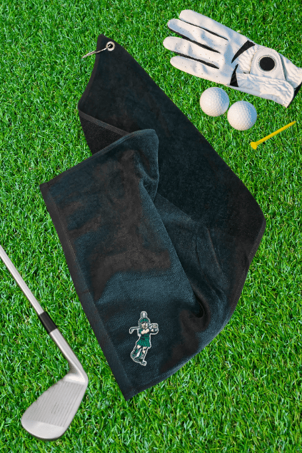 A black golf towel with a small embroidered golfer logo, laid out on green artificial grass alongside a golf club, golf glove, two golf balls, and a yellow tee.