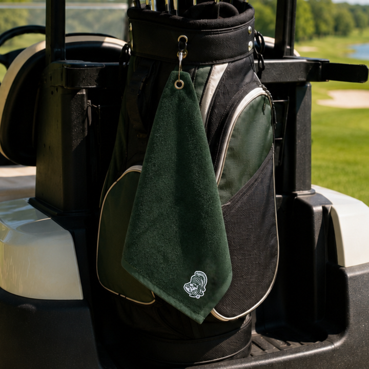 A dark green golf towel with a white embroidered logo hangs from a black and green golf bag on a golf cart, with a golf course background visible.