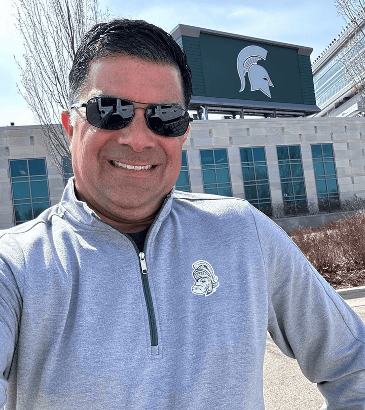 Michigan State Light Grey OGIO Quarter-zip with Gruff Sparty Logo on Male model in front of Spartan Stadium East Lansing