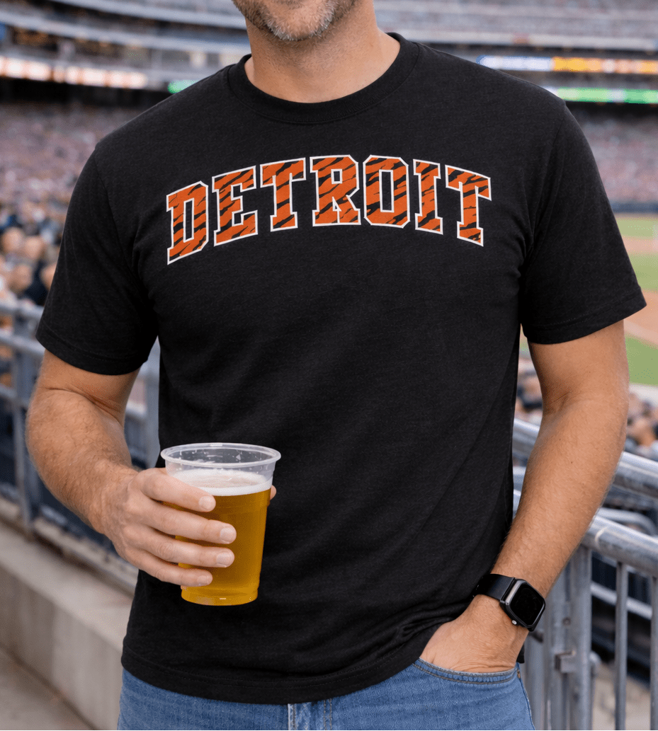 Male model wearing black shirt with Detroit logo with tiger stripes design holding a beer at a baseball stadium