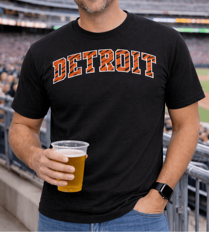 Male model wearing black shirt with Detroit logo with tiger stripes design holding a beer at a baseball stadium
