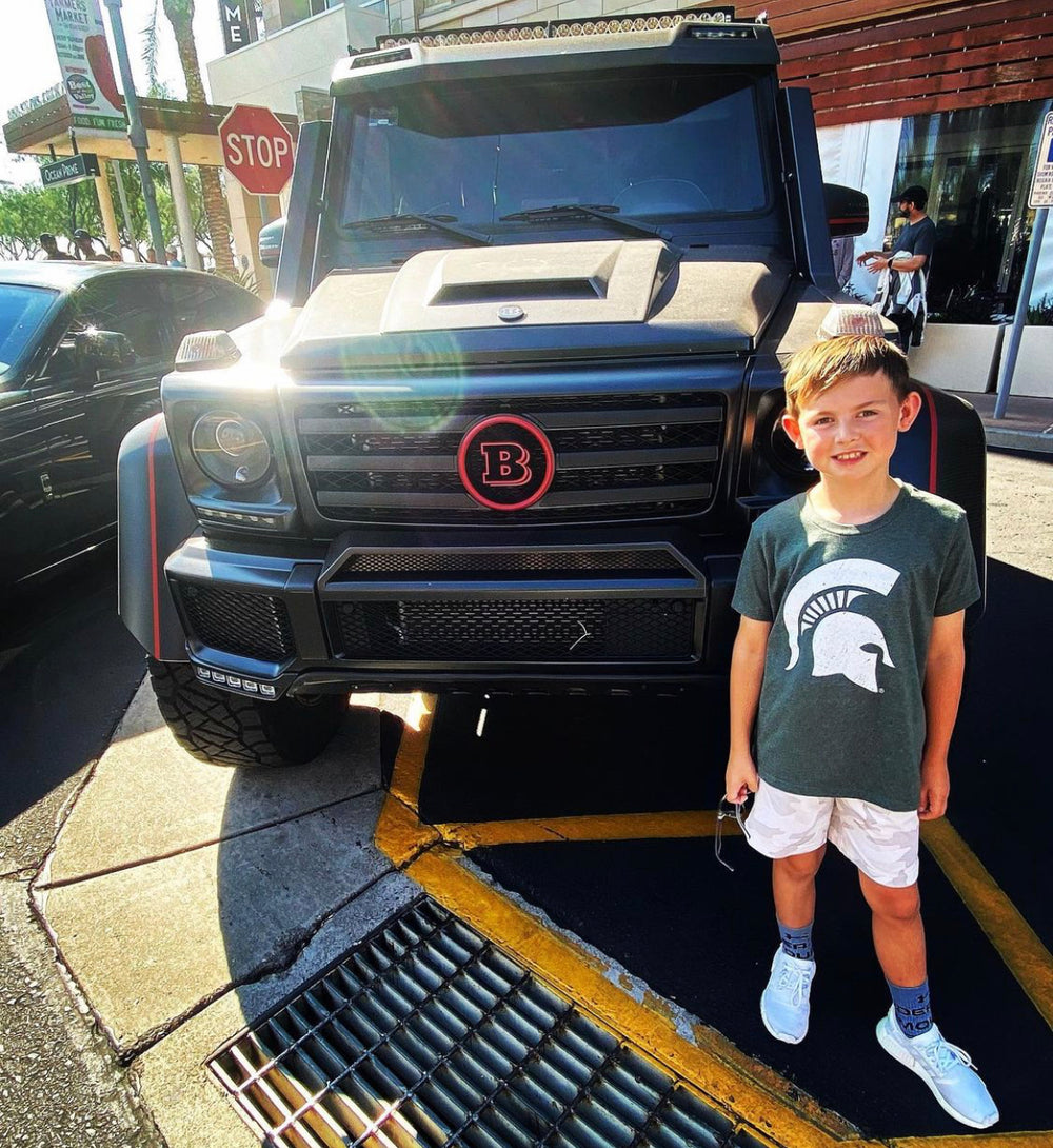 A young boy wearing a dark green Michigan State Spartans t-shirt stands in front of a black customized Mercedes-Benz G-Class vehicle. The vehicle features a prominent red circle badge with a white 'B' on the grille, indicating the brand is Mercedes-Benz.