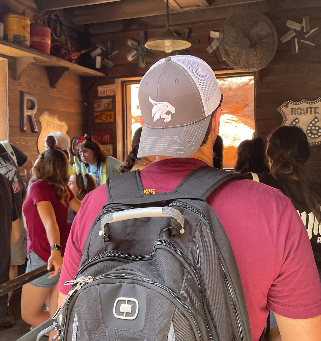 Model wearing backwards Texas State Bobcats Trucker hat that is grey in front and white mesh in back standing in line at Disneyland