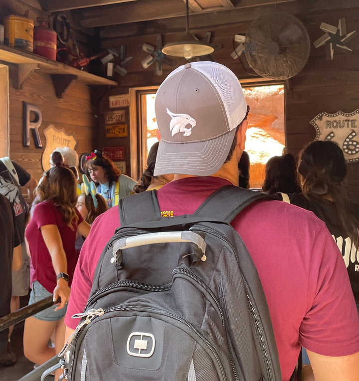 Model wearing backwards Texas State Bobcats Trucker hat that is grey in front and white mesh in back standing in line at Disneyland